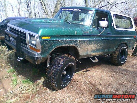 Green Ford Bronco with white top, lifted on mud tires, covered in mud, parked on dirt