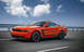 2012 Ford Mustang - A bright orange 2012 Ford Mustang Boss 302 is captured in a three-quarter front view while driving on a highway under a cloudy sky.