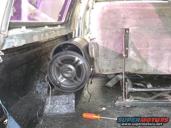 Interior of a vehicle showing a large speaker mounted on the floor, a worn seat, and a screwdriver