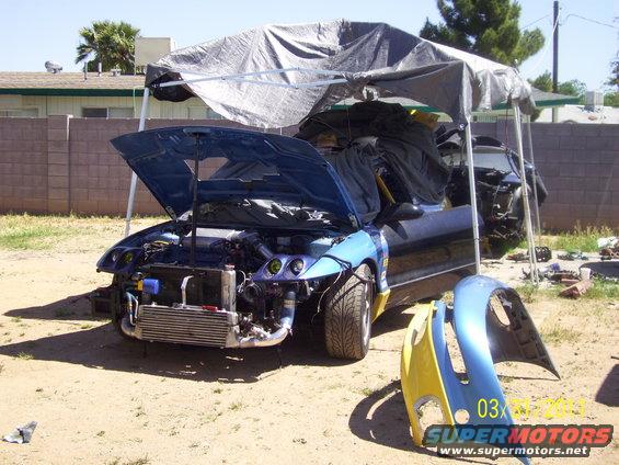 blue car with hood open, engine exposed, parts removed, parked under a tarp