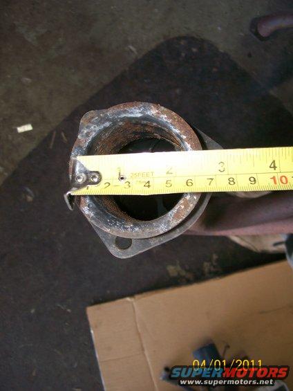 Measuring the diameter of a rusty exhaust pipe with a tape measure. The pipe has a flange at one end
