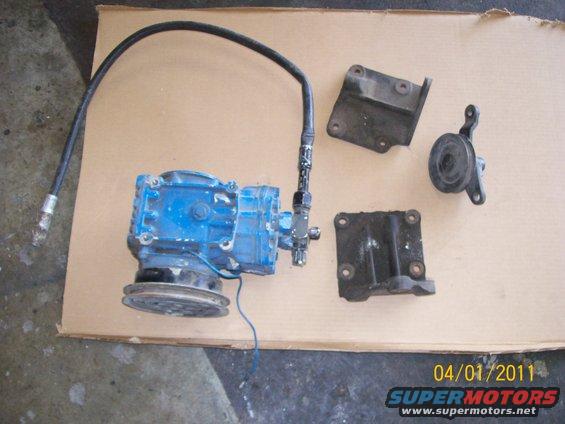 Blue air conditioning compressor with black hose and three metal mounting brackets on cardboard