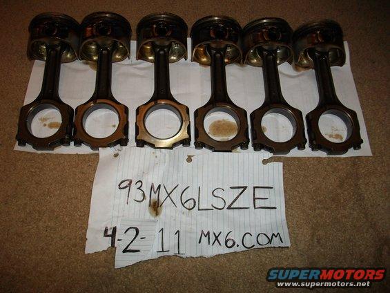 Six used engine connecting rods and pistons laid out on a carpeted surface