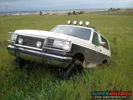 White and black 1989 Ford Bronco stuck in a grassy field with off-road lights