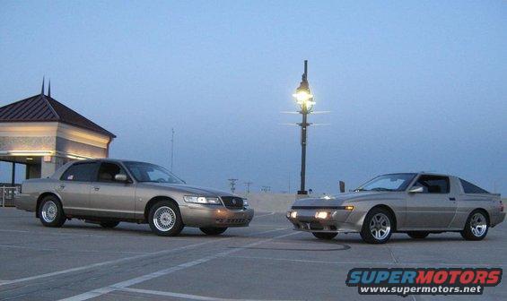 Silver 1980s coupe and a tan sedan parked on pavement, with a building and lamppost in the background
