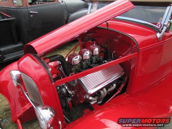Red hot rod with hood open showing V8 engine with triple carburetors and chrome valve covers. Black pickup truck in background
