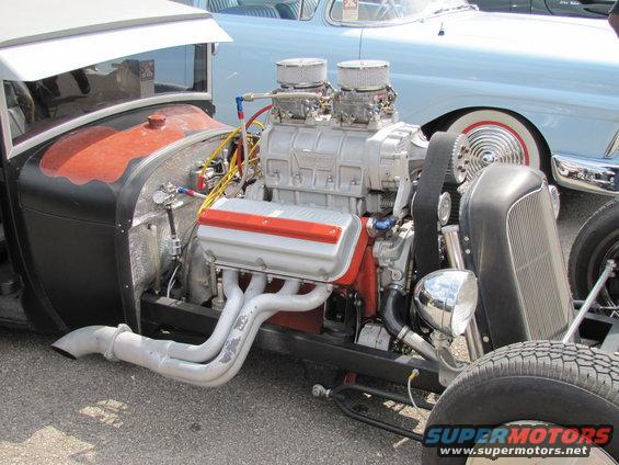 Hot rod engine with a supercharger and dual carburetors. A light blue classic car is visible behind it
