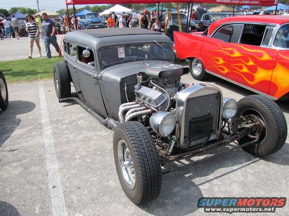 Gray hot rod sedan with exposed engine and side pipes, parked next to a red car with flames