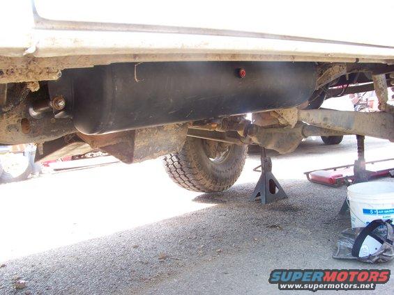 Undercarriage of a white vehicle showing a black fuel tank and suspension components, supported by jack stands