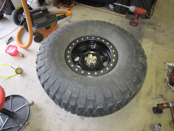 Large off-road tire on a black beadlock wheel with silver lug nuts, resting on a concrete floor
