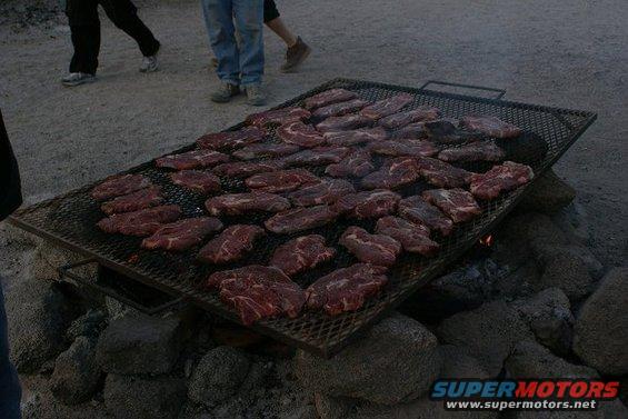 Many raw steaks cooking on a large metal grill over rocks and coals