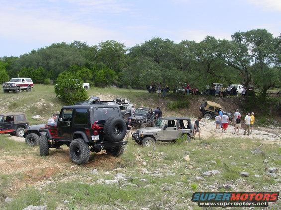 Several off-road vehicles parked on a rocky, grassy hill with trees in the background