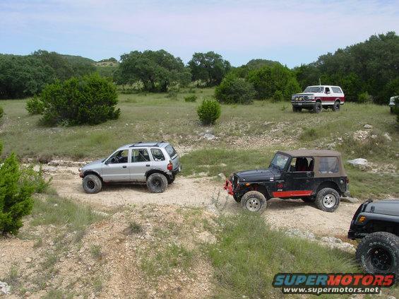 Several off-road vehicles, including a silver SUV and two Jeeps, on a dirt path in a grassy, wooded area