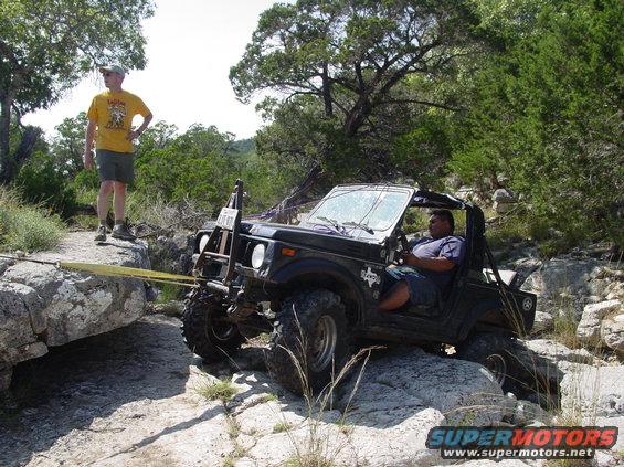 Black off-road vehicle with a man driving, being pulled by a yellow strap over rocks. Another man stands nearby