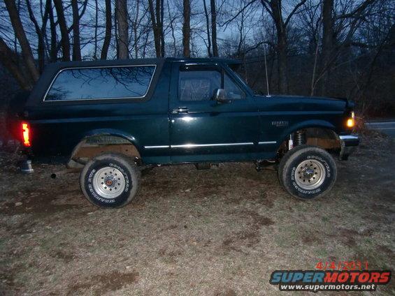 Side view of a dark green SUV with a soft top, lifted suspension, and aftermarket wheels parked on a dirt road