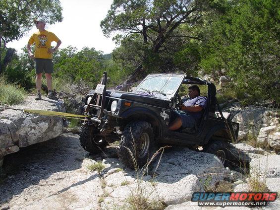 Black off-road vehicle with a man driving, on rocks, with a yellow tow strap attached to a large rock
