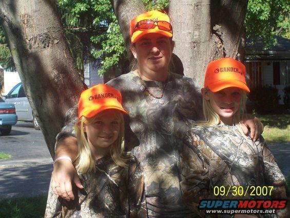 Three people in camouflage and orange hats stand near a tree. A blue car is partially visible on the left
