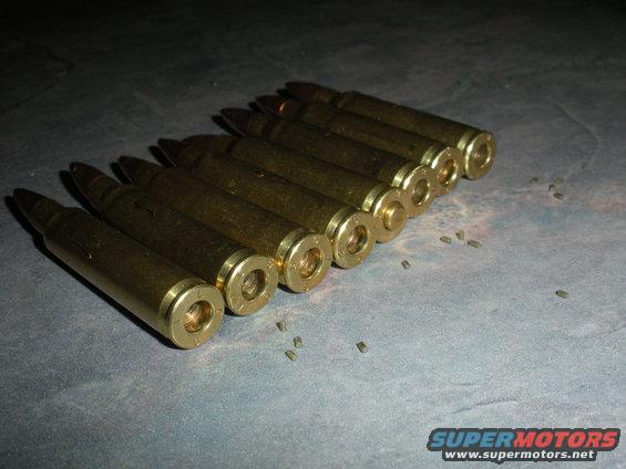 Row of brass cartridges on a gray surface with small debris scattered around. A Supermotors.net logo is in the lower right corner
