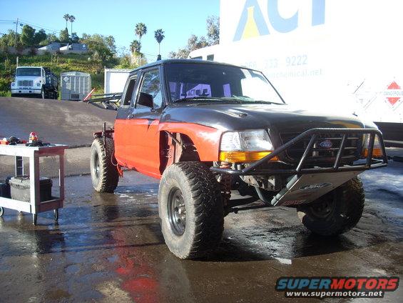 Modified pickup truck with orange fenders and a black hood. It has large off-road tires and a custom front bumper with a Ford logo
