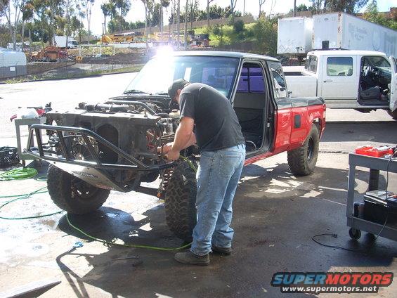 Man works on front end of red pickup truck with aftermarket bumper. Another white truck is parked behind it