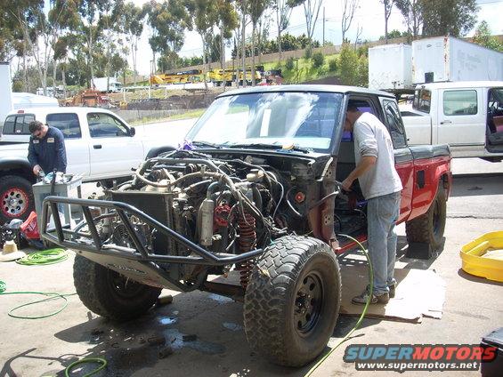Red pickup truck with front end removed, showing engine and off-road suspension