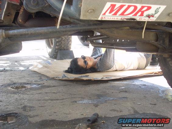 Person lying on cardboard working under the chassis of a vehicle; MDR sticker visible