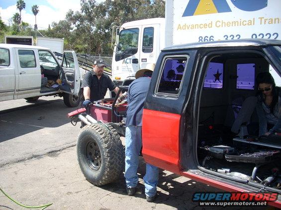 Two guys working on a red and black truck chassis. Another white truck is parked nearby