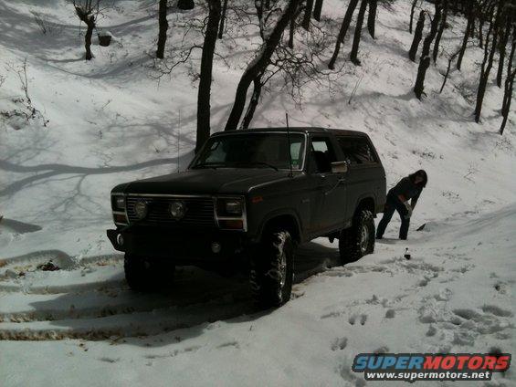 Dark colored Ford Bronco with aftermarket bumper and large tires parked in snow, person bending down near rear