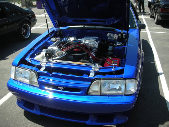 Blue Ford Mustang with hood open, showing a supercharged engine. Another black Mustang is parked to the left
