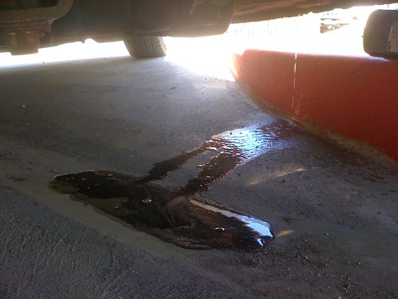 underneath car, dark fluid puddle on concrete, red drain pan nearby