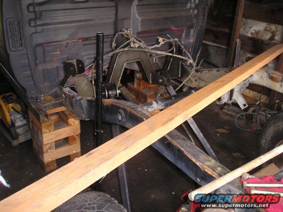 Truck frame on blocks with suspension components visible. A wooden plank lies across the frame