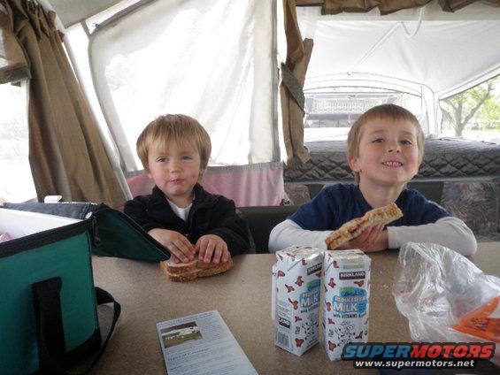 Two young boys sit at a table inside a pop-up camper, eating sandwiches and drinking milk