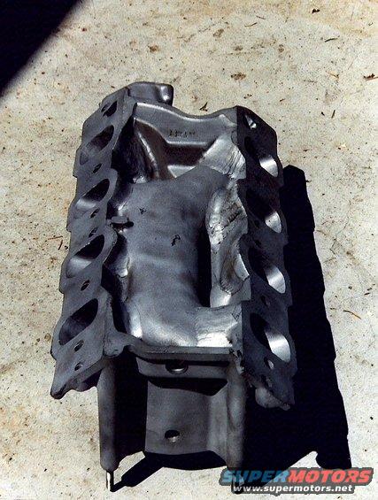 Engine block casting with visible port openings and internal structure, resting on a concrete surface