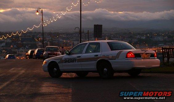 Sheriff's car parked on a street with a town and hills in the background. Lights strung overhead