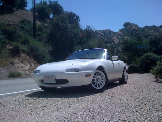 White 1991 Mazda Miata parked on a gravel shoulder next to a road with hills in the background