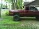 2001 Dodge 1500 - Maroon pickup truck with large tires parked in a grassy area near a wooden building.
