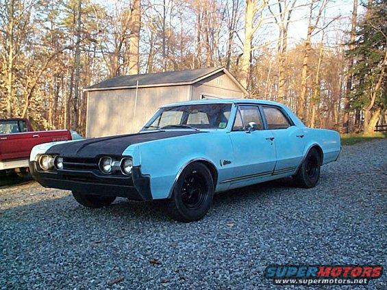 1966 Oldsmobile Cutlass sedan, light blue with black hood, parked on gravel lot