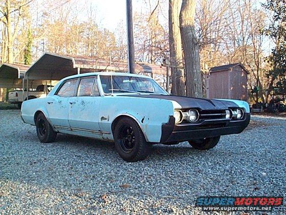 1966 Oldsmobile Cutlass sedan, light blue with black hood, parked on gravel, front 3/4 view