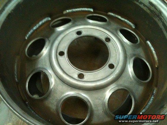 Close-up of a custom steel wheel with multiple lug holes and a central hub. Welds are visible around the rim