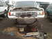 1986 Ford Bronco - A white Ford Bronco sits in a junkyard with its hood open and missing tires.