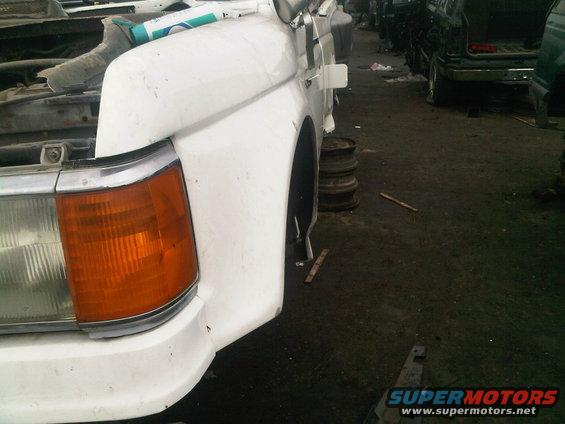 White pickup truck front fender, headlight, and grille area. Other vehicles visible in background