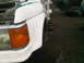 1986 Ford Bronco - Front corner of a white pickup truck in a junkyard setting with other vehicles around.
