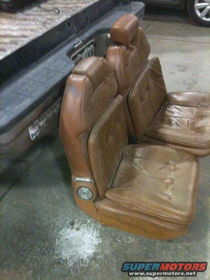 Brown leather bucket seats with headrests, one seat has a silver dial on the side. Part of a truck bed is visible behind them