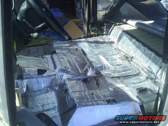 Vehicle interior floor covered in silver sound deadening material, showing exposed metal and some wiring
