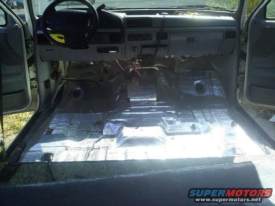 Interior of a vehicle with the carpet removed, showing sound deadening material on the floor and dashboard