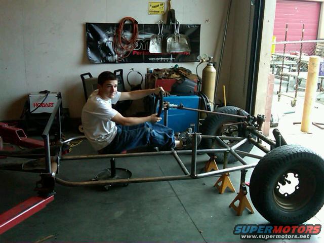 Tube frame chassis on jack stands with a person sitting in the driver's seat, holding a steering wheel