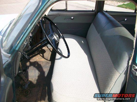 Interior of a 1950s car showing a light-colored bench seat, black steering wheel, and dark green dashboard