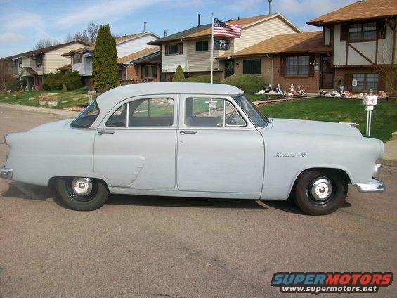 Gray 1952 Ford Mainline sedan parked on street, side view. Houses and American flag in background