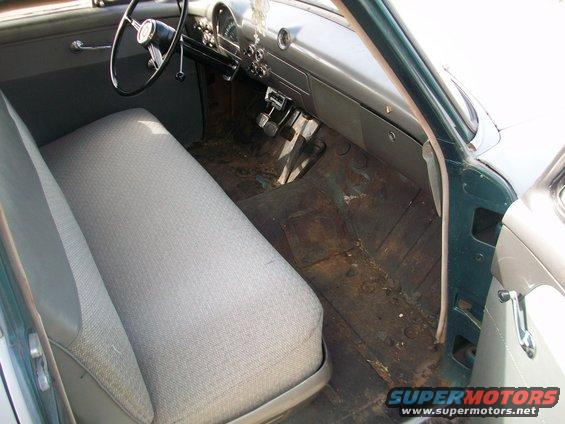 Car interior with grey patterned bench seat, dark dashboard, and floor pedals. The floor is rusty