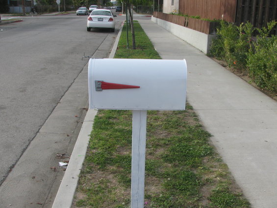 White mailbox with red flag on post next to sidewalk. White car parked on street